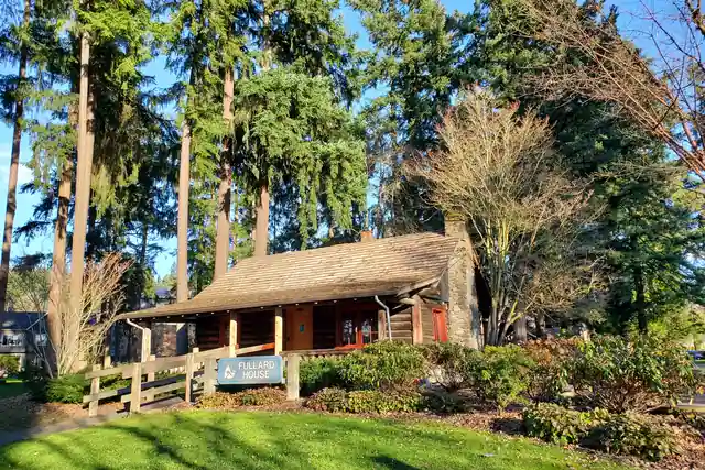 A low log cabin with a mossy shingled roof and a small porch sits among tall evergreens, with a green lawn in front.