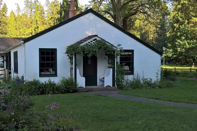 A small white farmhouse with dark green trim and a vine-covered porch stands on a lawn, with tall evergreens behind it.