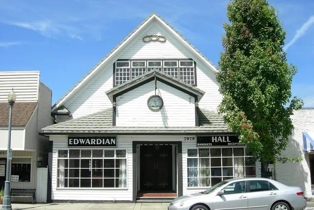 A white two-storey wooden hall with a steep gable and a round window, with a tall tree beside it.