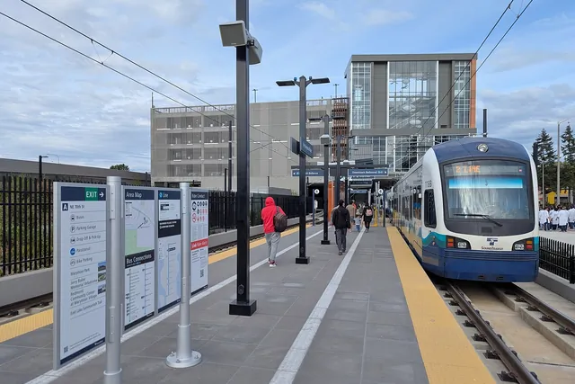 A blue and white light rail train stands at a new open-air platform with lamp posts and a glass stair tower behind.