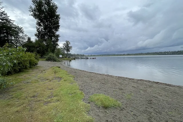 A curving gravel shoreline with a strip of grass and tall trees meets calm lake water under heavy clouds.