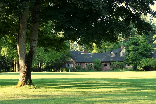 A long brown shingled house with tall chimneys behind a wide green lawn, framed by a large shade tree.